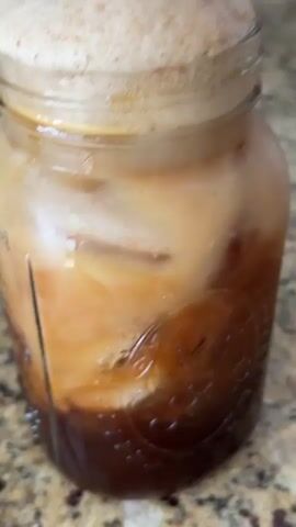 A close-up shot shows a mason jar filled with a layered beverage. At the bottom, there's a dark liquid, possibly coffee or espresso. Above this layer is a lighter, milky liquid, and ice cubes are also visible. Crowning the jar is a thick layer of tan-colored foam. A metal straw with a perforated end is inserted into the foam, causing it to bubble and froth. The jar sits on a speckled beige surface. A glimpse of a red and white patterned object appears briefly in the background. The lighting is bright, emphasizing the drink's textures and layering.