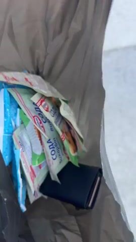 The video starts with a close-up view into a gray shopping bag, revealing multiple packets of Dr. Oetker baking soda, a blue packet of wet wipes, and a small navy blue wallet. The camera then pans up to show a street scene on a sunny day. Two cars, one gray and one white, are parked beside a tall building, which appears to be an office block or municipal building, topped with a communications tower. Green trees frame the scene.