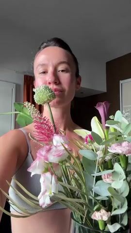 A woman with her eyes closed is smelling a bouquet of flowers. She then opens her eyes and smiles at the camera while holding the flowers. The woman is wearing a gray sports bra and white pants. She is standing in front of a window with orange curtains and a dresser in the background. The video is shot from the chest up, and the lighting is natural.