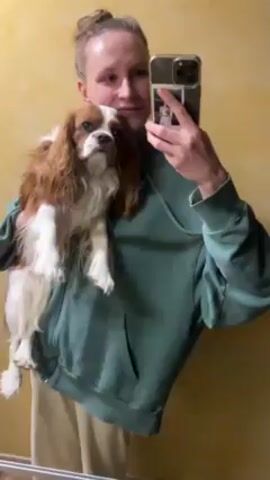 A woman holds a brown and white Cavalier King Charles Spaniel in front of her and takes a selfie in a bathroom mirror. The woman has light skin and her hair is pulled back in a bun. She is wearing a green hoodie and tan pants. The dog is facing forward with its ears hanging down. The bathroom wall is a yellow-orange color. The woman holds up her phone, which has a photo attached to the back, and points the camera towards the dog's face. The video ends in a blurry shot of the dog's face and the woman's hand holding the phone.