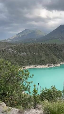 The video pans across a wide turquoise lake surrounded by rugged, green hills and mountains under a cloudy sky. The foreground features sparse vegetation including grasses and spiky plants. The camera movement captures the vastness of the landscape, showcasing the unique color of the water and the mountainous terrain.