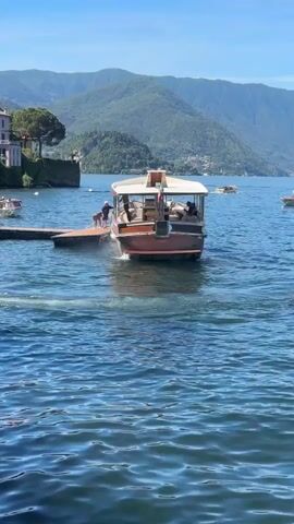 A wooden boat with a white canopy and passengers is docked at a wooden pier on a lake. The boat is gently bobbing in the water. Several people are on the pier and on the boat. In the background, there are green, tree-covered mountains and a few buildings with red tile roofs. The sky is clear and blue. The water is blue with white caps.