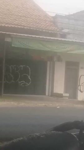 A hazy, overexposed video shows a storefront with a green awning. The awning has text on it, but it is difficult to read due to the lighting. There is graffiti on the metal doors of the storefront. To the right of the doors, there is a white wall with another door. The foreground is dark and appears to be the seat of a motorcycle, with some of its frame visible. The overall environment appears to be an outdoor urban or semi-urban setting, possibly on a cloudy or dusty day.
