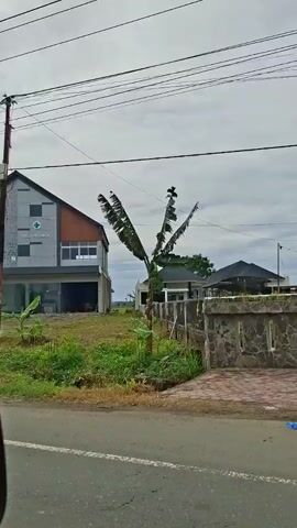 The video shows a roadside view of a two-story building under construction in a rural area. The building has a sign that says "VILA MEDIKA" with a green cross emblem.  The first floor has large glass windows. The second floor has a unique gray and white patterned design element and some regular windows. The building's roofline is dark gray. Next to the building is a large expanse of green grass. Behind the building are other buildings, a field, and a cloudy sky. The camera moves to show a bus stop with a thatched roof. The road in front of the building is paved and has a median of green grass.  Overhead are numerous power lines.