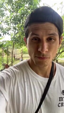 The video is a selfie-style recording of a man wearing a white t-shirt and a black baseball cap. He is standing outdoors, with a rustic outdoor oven or fireplace visible behind him. Inside the oven, a large pot is on a grate over a fire, with smoke billowing from it. The man is looking directly at the camera with a neutral expression. The environment appears to be a rural or tropical setting, with greenery visible in the background. The lighting suggests it is daytime.