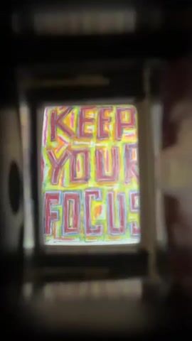The video shows a close-up view through a viewfinder or a small opening. In the center of the frame, a colorful sign with the words KEEP YOUR FOCUS is visible. The letters are handwritten and outlined in various colors, including red, yellow, and blue, against a light background. The background behind the sign appears to be white with some faint scribbles of color. The edges of the viewfinder are dark and blurred, framing the central image. The lighting appears to be artificial and somewhat dim, creating a vignette effect around the sign. There are no people or active interactions visible in the video.