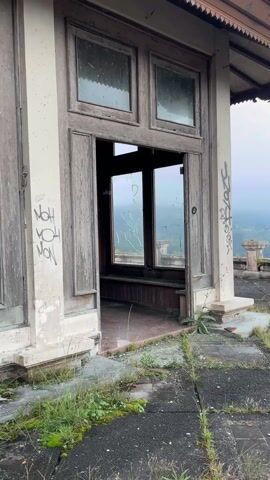The video shows an exterior view of a decaying building. The structure has old wooden doors and windows, many of which are broken. The exterior walls are discolored and marked with graffiti. Grass and weeds grow around the base of the building. Beyond the building, a mountainous landscape is visible under a cloudy sky.