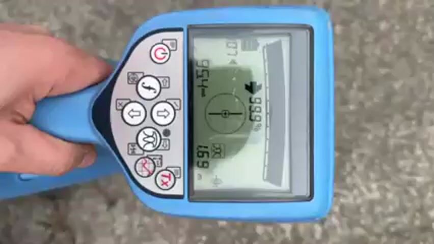The video shows a person using a utility locator device on a concrete surface. The device is light blue with a digital display showing a compass-like graphic, numerical data, and an arrow pointing upward. The screen also shows a battery indicator. The person's hand is visible holding the device, and their legs and boots are also visible. The device's display shows readings like "953 mA" and "99.9%." As the person moves, the readings on the display change. The camera angle alternates between a close-up of the device's screen and a wider shot showing the person's feet and the surrounding concrete. A yellow spray paint can is briefly visible.