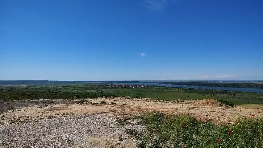 The video shows a wide panoramic view of a flat landscape under a bright blue sky. A wide river flows through the middle ground, with green fields and vegetation on both sides. In the distance, there appears to be a town or settlement. The foreground consists of a dirt and gravel area with patches of green grass and clusters of red poppy-like flowers. The camera is static, and the overall impression is of a peaceful and rural scene.