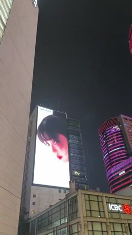 The video begins with a close-up of a large LED screen on the side of a building, displaying a woman's face with striking red lipstick and dark bangs, bathed in red lighting. The camera then pans up and to the right, revealing more of the cityscape at night. Several skyscrapers dominate the frame. The initial building with the LED screen has a vertical layout. To its right, another building features a display that transitions from the woman's face to her upper body, wearing a white top and a red skirt. Further to the right, two more skyscrapers are illuminated with vibrant purple lights, creating a dazzling effect. The camera continues to pan, showing the sheer scale of these illuminated structures against the dark night sky. The base of the buildings reveals commercial signage, including "ICBC" with its red circular logo, indicating a financial institution. Green foliage is visible in the foreground, partially obscuring the lower parts of the buildings. The overall scene captures a dynamic urban environment at night, characterized by large-scale digital displays and colorful architectural lighting.