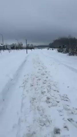 The video shows a snowy path leading away from the camera. The path is covered in snow that has been churned up by footprints and tire tracks. On either side of the path is a deeper layer of untracked snow. In the background, there are streetlights lining the path, and some buildings can be seen on the left. On the right is a line of trees. The sky is overcast.