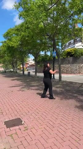 A woman wearing a black outfit and sunglasses stands on a red brick path, appearing to speak to someone or something up in a tree to her right. The tree is leafy and green. Beyond the tree is a street with a pedestrian crossing sign, as well as buildings in the background. She gestures with her hands, and the sky is blue with some clouds.