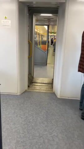The video shows an open sliding door between two train cars. The interior of the next car is visible, including rows of seats that alternate between orange and blue. Yellow handrails are attached to the backs of the seats. A few people can be seen at the far end of the car. The train car is well-lit by overhead lights. The camera is stationary and the train appears to be in motion.