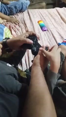 A close-up video shot indoors shows an adult and a child on a bed, both holding a black Playstation controller. The adult’s hands grip the controller while the child’s smaller hand is visible near the buttons, appearing to be interacting with the controller as well. Other children are on the bed in the background. A colorful toy is placed on the pink patterned bed sheet. The adult wears a dark-colored shirt, and the child is wearing grey clothing with camouflage pants. The audio appears to feature music.