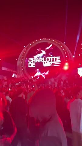 The video shows a concert or festival at night. A large circular screen displays the words "CHARLOTTE DE WITTE" in large white letters, with a pink animated character in the center. Below the screen, a crowd of people are visible, many with their hands raised. Red and white lights flash across the scene. The camera is positioned behind a person wearing a dark hooded sweatshirt, who is looking towards the stage. The lighting shifts from white and blue to predominantly red, creating a dynamic and energetic atmosphere.