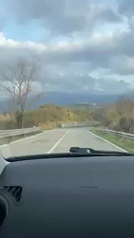 The view from inside a car shows a winding asphalt road stretching into the distance, flanked by grassy hills and scattered vegetation. Metal guardrails line the edges of the road. In the background, a mountain range is visible beneath a sky filled with heavy, overcast clouds. Music is playing in the car. The camera appears to be mounted on the dashboard of the car.
