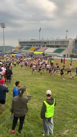 This media file captures a large running race in progress, set against the backdrop of a stadium with tiered seating. The subjects are numerous people, primarily participants in athletic wear, actively running across a grassy field. Spectators are also visible, some holding up phones to record the event.

The activity taking place is a race, with runners in various positions, some at the front of the pack and others further back. The stadium suggests this is a formal event, possibly a marathon or a significant race.

The time of day appears to be daytime, with overcast skies. The weather seems cloudy, but not actively raining. The location is identifiable as Talgar, Kazakhstan, indicated by text on a runner's shirt that reads "ALMATY" (a major city in Kazakhstan, near Talgar).

Notable details include the presence of a volunteer wearing a high-visibility vest with text that appears to read "VOLUNTEER" and possibly further details like "RACE." The atmosphere seems energetic, with many people participating and observing. The finish line is visible in the distance, adorned with banners.