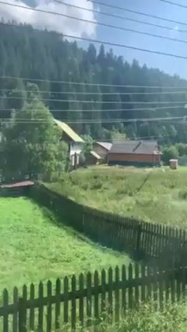 The video shows a view from a moving train. The scenery consists of green fields, wooden houses, fences, power lines, and a forest-covered hill in the background. The sky is blue with some clouds. The scene appears to be a rural or mountainous area.