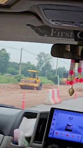 This is a first-person view from inside a vehicle looking out through the windshield. The vehicle appears to be moving on a road under construction. In the foreground, a dashboard display shows a speed of 82 km/h, which decreases to 80 km/h and then 79 km/h as the video progresses. The road ahead is partially paved and partially dirt. Several construction vehicles are visible, including a yellow roller on the left and a yellow grader in the distance on the right. Orange traffic cones and temporary white and pink barriers line the road. The sky is overcast. A decorative string of pink and yellow tassels hangs from the rearview mirror.