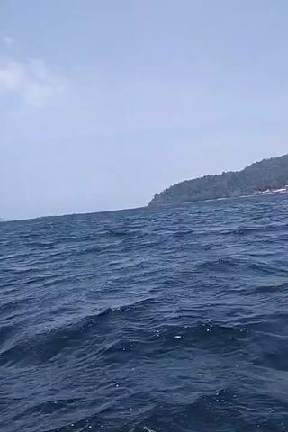 The video shows a choppy, dark blue ocean under a cloudy sky. In the distance, a treed island is visible on the right side of the frame. The camera appears to be on a boat, as evidenced by the movement of the water. There are no people or other objects clearly visible in the frame.