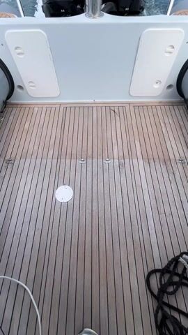 The video shows the back deck of a motorboat moving through water. The boat has a wooden deck with dark seams, and two black Mercury outboard motors. There is a stainless steel bar separating the deck and motors. The boat is gray with inflated, gray pontoons. The sky is blue with white clouds and land is visible in the distance. The boat is creating white water as it moves. There are ropes laying on the deck.