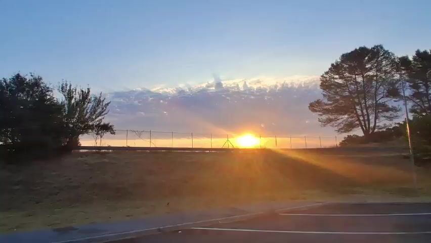 The video shows a sunrise over a hilly landscape. The sun is low on the horizon, casting a warm, golden glow. Rays of sunlight stream through the clouds, creating a dramatic effect. A fence runs across the middle of the frame, separating the foreground from the background. Trees and bushes are visible on either side of the fence. The foreground is a road or parking lot.