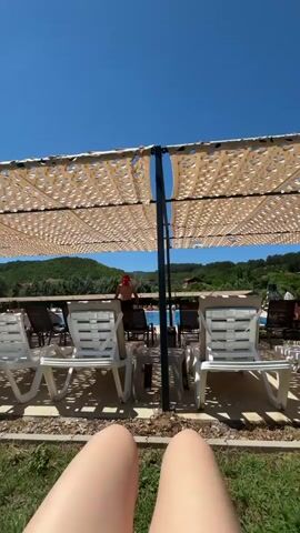 The video shows a first-person perspective of someone sitting on grass in front of two empty white lounge chairs. Behind the chairs is a shaded area with a tan, perforated canopy supported by a dark pole. In the background, there is a swimming pool with people in and around it, as well as more lounge chairs and trees on rolling hills under a clear blue sky. The overall scene depicts a sunny day at an outdoor pool or resort.