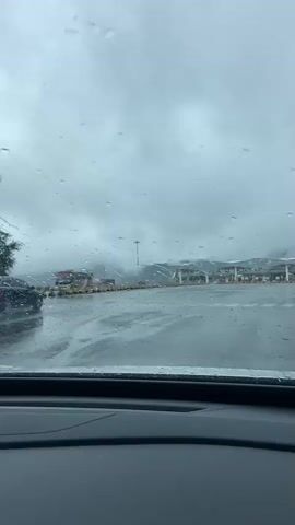 The video is shot from inside a vehicle, looking through a rain-streaked windshield. Several cars are visible on a wet road, with one black car in the left lane appearing to be the closest. Beyond the cars, a toll plaza or service area is visible in the distance, with multiple booths and awnings. The sky is overcast and appears to be raining heavily, with raindrops visible on the windshield. The overall scene suggests a view from a car driving towards or stopped at a toll station in rainy weather.