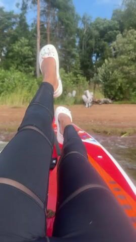 The video shows a first-person perspective of someone sitting on a paddleboard on a body of water. The person is wearing black leggings with mesh paneling and white slip-on shoes adorned with rhinestones. Their legs are extended forward, with one leg bent at the knee and the other resting on the paddleboard. The paddleboard is red and white with colorful graphics. In the background, there is a sandy shore lined with trees and vegetation under a bright, sunny sky. The water appears calm with small ripples.