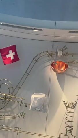 The video shows the interior of a Lindt Chocolate store, focusing on a kinetic sculpture display related to chocolate making. The display features a winding track made of gold-colored metal, incorporating various elements like a copper bowl, a chef's hat, a spiral ramp, and rotating gears. Golden balls travel along the track. A large screen displays a chef in a white uniform and chef's hat. People are visible in the background, including a woman with curly hair taking photos of the display with her smartphone. The walls are white and curved, with the Lindt logo displayed. The lighting is bright and even.
