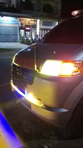 The video shows the front and side of a white ambulance parked on a dirt road at night. The ambulance has its headlights and emergency lights on. The emergency lights are red and appear to be a light bar on the roof of the vehicle. A red stripe runs along the bottom of the ambulance, and there is a red cross emblem on the side. The word "AMBULANCE" is written in red on the side of the vehicle.

In the background, there are buildings with lights on, and streetlights. A person wearing a hat and dark clothing is standing near the ambulance, appearing to be smoking. The scene is dimly lit by the ambulance's lights and the distant streetlights, suggesting it is nighttime. The ground is covered in dirt and gravel, with a few plastic bottles scattered on it. The overall impression is of an operational ambulance on standby or at a scene in a semi-urban or rural area. There is text visible on a building in the background that reads "SWISS BARBERSHOP".