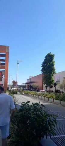 The video shows a sunny day outdoors in front of a modern shopping center. A person in a white shirt walks towards the left of the frame in the foreground. Behind them, a metallic walkway or grate is visible. In the mid-ground, a row of planter boxes with green and yellow foliage runs along a paved walkway. Beyond that, the shopping center building is visible with its beige and orange facade, a tall green tree, and other smaller trees. People can be seen standing and walking in front of the shopping center. A large white building or apartment complex is visible in the background on the right, and another similar building is partially visible on the left. A street lamp stands on the left side of the frame. Signs for "DECATHLON" and "FORUM" are visible on the building. The sky is a clear blue.