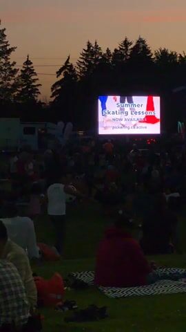 A large outdoor screen displays advertisements for "Summer Skating Lessons" and "Free Teen Stuff." A crowd of people sits on blankets and in chairs on a grassy field, watching the screen. In the background, trees and a stage with a band are visible. The sky is a gradient of orange and purple, indicating dusk. Several portable toilets and RVs are parked on the left side of the field.