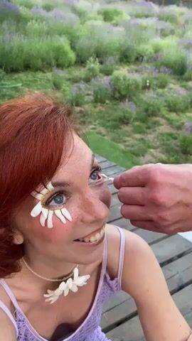 A woman with red hair and blue eyes is sitting on a wooden deck. She has daisies applied to her face as makeup, with some petals on her eyelids and cheeks. She is wearing a purple top and a necklace with a daisy design. In the background, there is a field of purple flowers, possibly lavender, and some green foliage. A hand enters the frame from the right and appears to be adjusting a daisy petal on the woman's face.