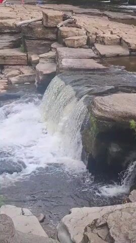 The video shows a small waterfall cascading over a series of flat, light-colored rocks. The water is white and frothy as it plunges into a darker, calmer pool below. The surrounding area is also rocky, with large, irregular stone formations that appear to be part of the riverbed. There are patches of green vegetation growing on some of the rocks, suggesting a natural, outdoor setting. In the distance, on the upper bank, several people can be seen, though they are small and indistinct. The lighting suggests it is daytime, possibly late morning or afternoon, with diffused light indicating it could be overcast or that the sun is not directly overhead. The overall impression is a peaceful, natural scene of flowing water and rugged, geological formations. The location is identified as Richmond, United Kingdom. There is no visible text in the media file.