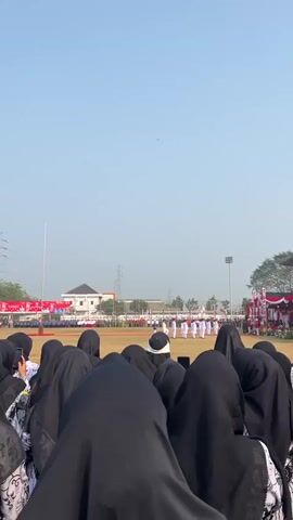 The video shows a crowd of people viewed from behind, all wearing black hijabs, facing a staged event on a field. The event features a line of individuals in white uniforms with dark head coverings, possibly a performance or ceremony, on a grassy field. In the background, there are tents with red and white decorations, buildings, and utility poles under a clear blue sky. The lighting suggests it is daytime.
