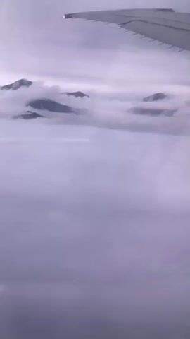 A view from an airplane window shows a wingtip in the upper right corner. Below the wing, a vast expanse of clouds stretches out, obscuring the ground. Several mountain peaks emerge from the cloud layer, appearing as dark silhouettes against the hazy sky. The overall lighting suggests it is either dawn or dusk, or a heavily overcast day. The video is a static shot, with no discernible movement or action.