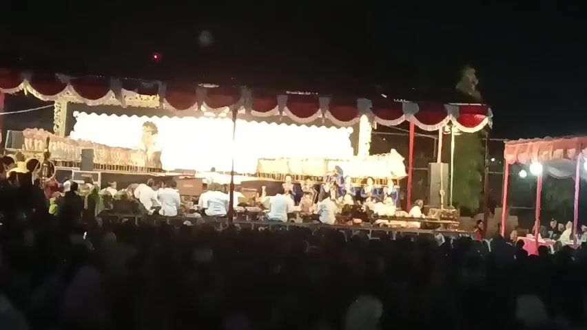A night-time outdoor performance takes place on a raised stage illuminated by bright lights. The stage is covered by a red and white striped canopy. On the stage, a group of musicians are seated, playing instruments. In front of the musicians, a white screen is lit from behind, suggesting a shadow puppet performance or a projection. A large crowd of people is gathered in front of the stage, watching the performance. The audience is largely out of focus and silhouetted against the stage lights. The environment appears to be a festival or cultural event.
