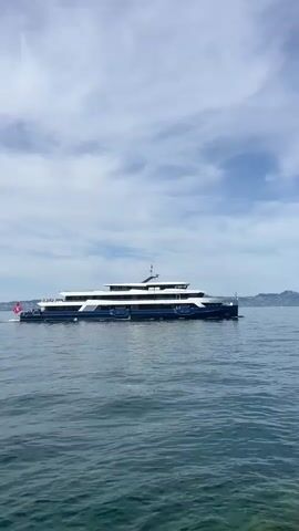 A wide shot captures a large, multi-decked navy blue and white passenger ship sailing across a body of water. The ship, identified by a Swiss flag at its stern, is moving from left to right across the frame. The water is a deep blue-green with visible ripples, and the sky above is a mix of blue and white clouds. In the distance, faint outlines of hills or mountains are visible on the horizon. The foreground shows a rocky shoreline. The video is a single, static shot with no discernible action other than the ship's movement.