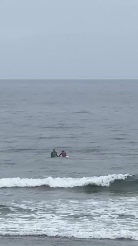Two people on surfboards are in the ocean. The person on the left is wearing a green wetsuit and the person on the right is wearing a pink wetsuit. They are facing each other and appear to be interacting. The water is choppy with small waves. The sky is overcast.