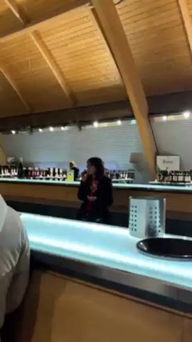 A woman standing at a bar is holding a wine glass, while someone else is partly visible to her left. The bar is long and illuminated from below. Behind the bar are rows of wine bottles. The woman is wearing a red shirt and a dark blazer. She has dark hair and glasses. A stainless steel utensil holder is visible in the foreground on the right side of the bar. The room has a high, sloped ceiling with exposed wooden beams. There are light fixtures suspended from the ceiling.