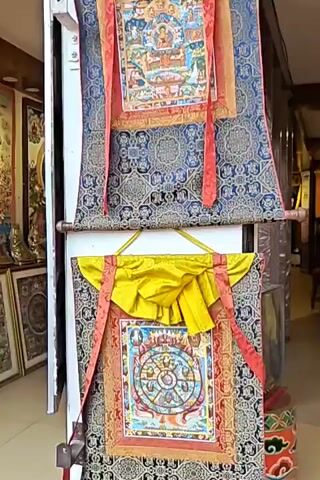 The video shows a close-up view of a display of Tibetan artwork and religious artifacts in a shop. The items include several thangkas (scroll paintings), some of which are elaborately decorated with gold and vibrant colors, featuring Buddhist imagery such as the Wheel of Life and various deities. The thangkas are adorned with red and yellow fabric tassels and streamers. In the background, other artworks, statues, and merchandise are visible, suggesting a retail environment focused on Tibetan culture and spirituality. The lighting appears to be artificial, illuminating the items for display. The camera slowly pans across the scene, providing a detailed look at the intricate craftsmanship of the art.