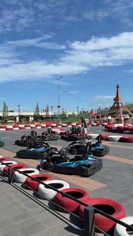 The video shows a go-kart track on a sunny day. Several go-karts are parked on the track, which is lined with red and white tires. In the background, there is an amusement park with rides, including a roller coaster and a Ferris wheel. There is also a replica of the Eiffel Tower. A person is walking on the track. The sky is blue with some clouds.