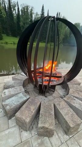 A wide shot shows a metal fire pit with burning logs. The fire pit is surrounded by large, rough-cut stones arranged in a circular pattern on a paved patio. Beyond the patio is a body of water, likely a lake or river, with a forest of evergreen trees on the opposite bank. The sky is overcast and appears to be either early morning or late evening. No people are visible in the video. The camera slowly zooms out, giving a broader view of the serene natural setting.