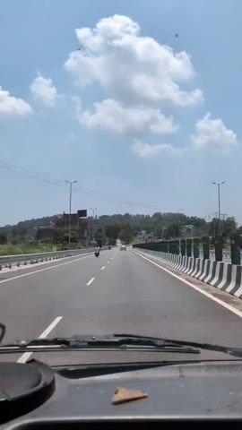 The video shows a view from inside a vehicle driving on a highway. The road is paved and has lane markings. There are guardrails and poles along the side of the road. In the distance, there are hills with some buildings and trees. The sky is blue with some white clouds. A few vehicles, including a motorcycle and a car, are visible on the road. The dashboard of the vehicle is partially visible in the foreground.