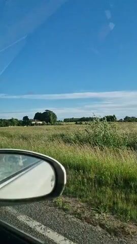 The video shows a view from a moving vehicle, looking out at a rural landscape. In the foreground, the side-view mirror of the vehicle is visible on the left. The landscape consists of green fields, tall grass, trees, and a house in the distance. The sky is clear and blue with a few white clouds. The overall impression is a peaceful, sunny day in the countryside.