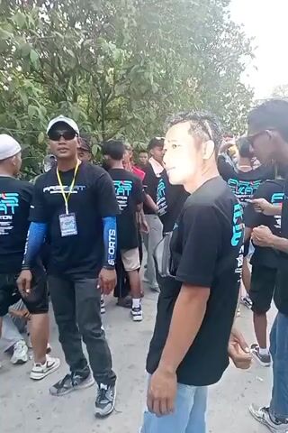 The video shows a group of young men outdoors, possibly at an event or gathering. Many of them are wearing black t-shirts with a logo that appears to be "ARS BEAT SPORTS." One man in the foreground, wearing a black t-shirt, dark pants, a cap, and sunglasses, is speaking and gesturing. He has a lanyard with an ID badge around his neck. Other individuals are standing and sitting around him, some looking towards the camera. The environment appears to be a street or pathway lined with trees and foliage. Some people are holding what appear to be cigarettes. The overall atmosphere seems casual and energetic.