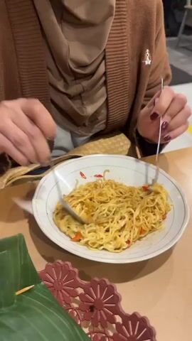 A close-up shot shows a person wearing a brown cardigan and a tan hijab, using a fork and spoon to mix a plate of instant noodles. The noodles are golden brown and appear to be seasoned with chili flakes and other spices. The plate is white with a gold rim, and it rests on a light brown table. In the foreground, there is a green banana leaf with a toothpick. The background is blurred, but hints of a dimly lit indoor or outdoor dining area are visible. The person's hands, with burgundy nail polish, are actively engaged in mixing the noodles, indicating they are preparing to eat or are in the process of eating.