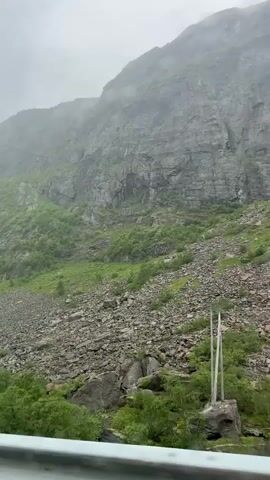 This video captures a scenic view of a rugged mountain landscape in Skare, Norway. The scene is dominated by a massive, steep, rocky mountain face, partially obscured by mist and clouds, suggesting overcast or rainy weather. The lower slopes of the mountain are covered in lush green vegetation, with patches of trees and smaller shrubs dotting the landscape. A significant scree slope, composed of countless loose rocks and boulders, runs across the mountainside, indicating past geological activity.

In the foreground, the lush green canopy of trees creates a vibrant contrast with the muted tones of the mountain and sky. A guardrail is visible at the very bottom of the frame, implying the footage is likely taken from a vehicle or a viewpoint along a road.

The overall impression is one of raw, natural beauty and dramatic geological formations. The misty conditions add an atmospheric and slightly mysterious quality to the scene, hinting at a cool, damp day. The time of day is not explicitly clear due to the overcast conditions, but the light suggests it is daytime. There is no visible text in the video. The scene evokes a sense of awe at the power and grandeur of nature.