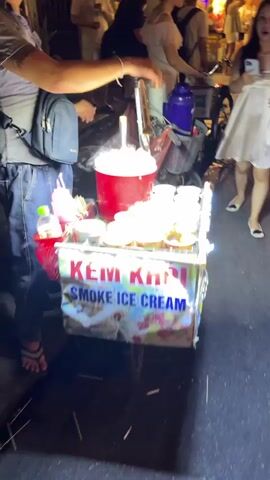 The video shows a street vendor selling "smoke ice cream". The vendor is using a large red container filled with a white substance that is producing smoke. Around the container, there are small cups filled with colorful candy balls. A person is holding a metal tool over the red container, presumably to create the smoke effect. In the background, other people are walking by, and there is a bicycle with a basket. The scene appears to be at night, indicated by the lighting and the presence of a street. The vendor's cart has a sign that reads "KEM KHOI SMOKE ICE CREAM".